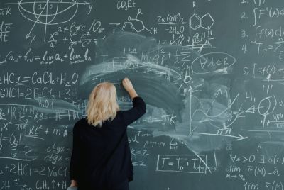 blonde woman writes complex formulas on a large chalkboard